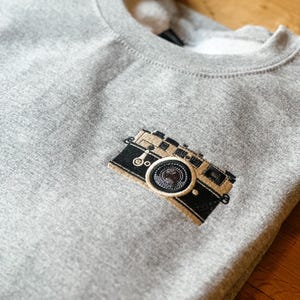 May include: A heather gray sweatshirt with a detailed embroidered camera design on the front. The camera is black and tan with intricate stitching, suggesting a vintage or retro style. The sweatshirt has a crew neck.