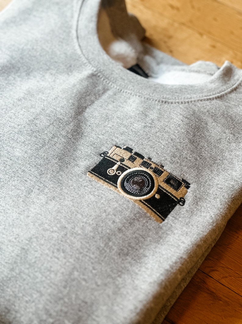 May include: A heather gray sweatshirt with a detailed embroidered camera design on the front. The camera is black and tan with intricate stitching, suggesting a vintage or retro style. The sweatshirt has a crew neck.