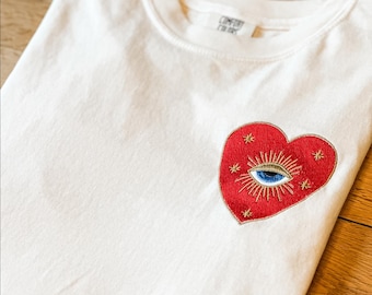 May include: A white short-sleeved t-shirt featuring an embroidered red heart patch on the left chest. The heart contains a central blue eye design surrounded by gold-colored radiating lines and small star accents. The shirt has a crew neckline.