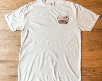 May include: A white short-sleeve crew neck t-shirt featuring a small graphic on the upper left chest. The graphic is a brown and tan retro-style sign shape containing a small pine tree icon and the text Protect Our PARKS in dark brown.
