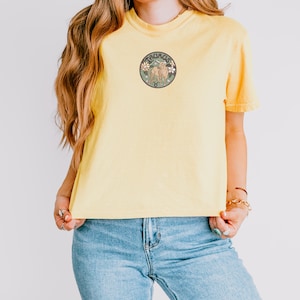 May include: A yellow short-sleeved t-shirt featuring a circular graphic on the chest. The design includes a brown bull illustration, the word TAURUS, and floral accents in shades of pink, white, and green against a dark background.