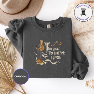 Puede incluir: Una sudadera de cuello redondo gris carb&oacute;n con un bordado de estilo medieval que muestra a una persona con t&uacute;nica y el texto "Thar goes! The last fvck I giveth". El dise&ntilde;o incluye adornos decorativos en tonos dorados y crema.