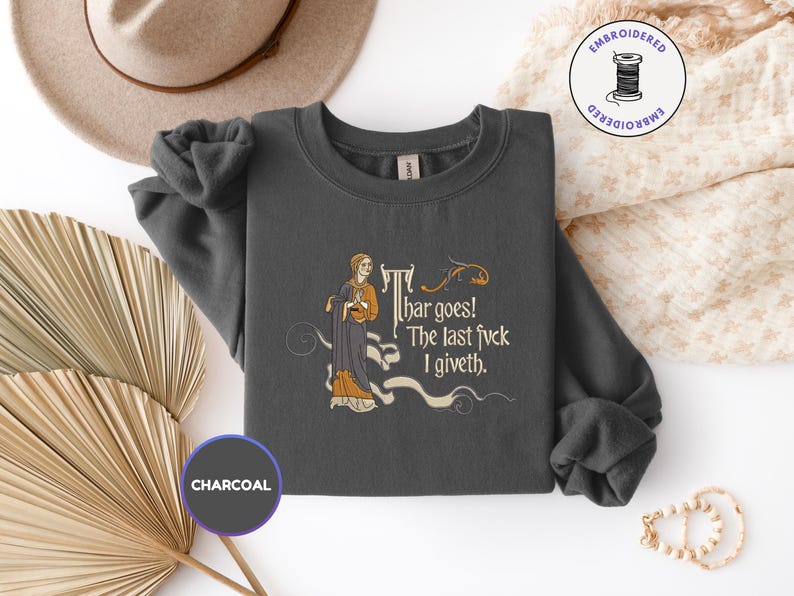 Puede incluir: Una sudadera de cuello redondo gris carb&oacute;n con un bordado de estilo medieval que muestra a una persona con t&uacute;nica y el texto "Thar goes! The last fvck I giveth". El dise&ntilde;o incluye adornos decorativos en tonos dorados y crema.