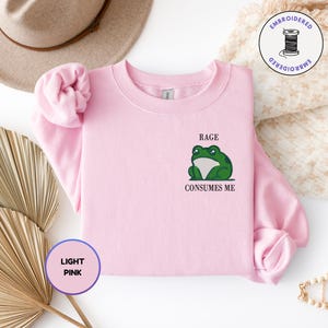 Pu&ograve; includere: Una felpa girocollo rosa chiaro con un ricamo raffigurante una rana verde sul petto sinistro. Sopra la rana &egrave; presente il testo RAGE e sotto la rana il testo CONSUMES ME. La felpa &egrave; dotata di maniche lunghe e di un colletto a coste.