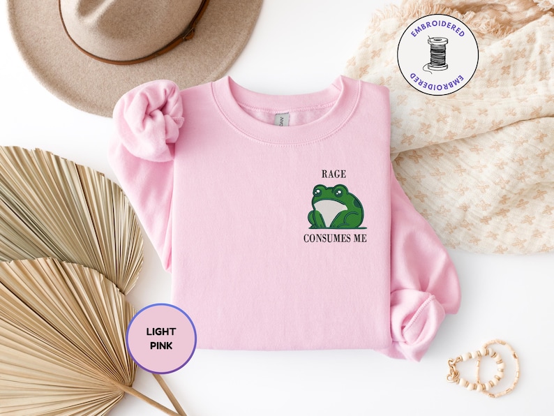 Pu&ograve; includere: Una felpa girocollo rosa chiaro con un ricamo raffigurante una rana verde sul petto sinistro. Sopra la rana &egrave; presente il testo RAGE e sotto la rana il testo CONSUMES ME. La felpa &egrave; dotata di maniche lunghe e di un colletto a coste.