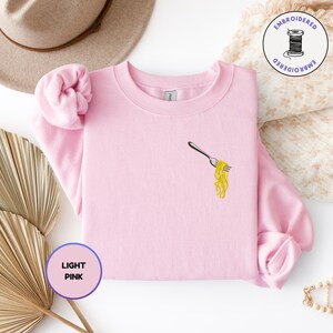 May include: Light pink sweatshirt with an embroidered design of a fork holding yellow spaghetti. The sweatshirt has a crew neck and long sleeves. The words "LIGHT PINK" are in a purple circle.