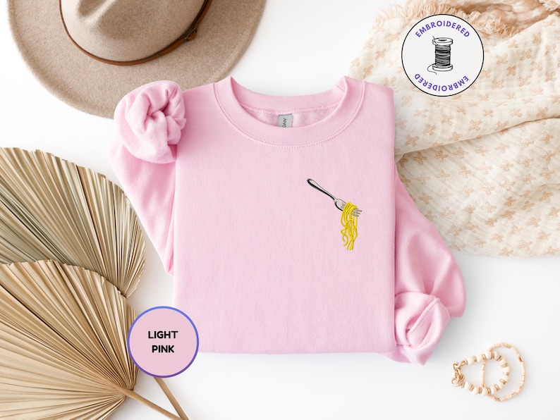 May include: Light pink sweatshirt with an embroidered design of a fork holding yellow spaghetti. The sweatshirt has a crew neck and long sleeves. The words "LIGHT PINK" are in a purple circle.