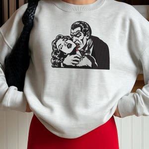 May include: A light gray crewneck sweatshirt featuring a black and white graphic print of a vampire biting a person's neck. The illustration shows a man in a suit with fangs and a drop of red blood on the neck of the person being bitten.