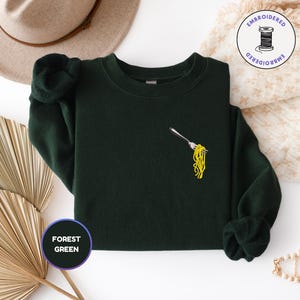 May include: Forest green sweatshirt with an embroidered fork holding yellow spaghetti. The sweatshirt has long sleeves and a crew neck. A round sticker with the words "FOREST GREEN" is in the lower left corner.