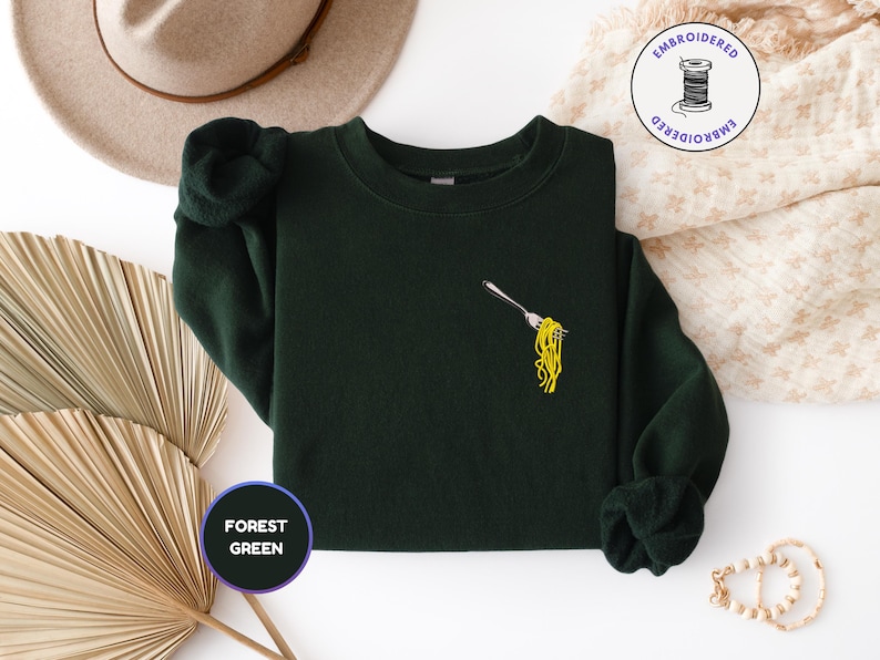 May include: Forest green sweatshirt with an embroidered fork holding yellow spaghetti. The sweatshirt has long sleeves and a crew neck. A round sticker with the words "FOREST GREEN" is in the lower left corner.