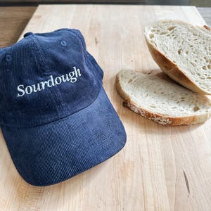 Può includere: Un berretto da baseball in velluto a coste blu con la parola "Sourdough" ricamata in bianco sul davanti. Due fette di pane bianco sono su una superficie di legno.