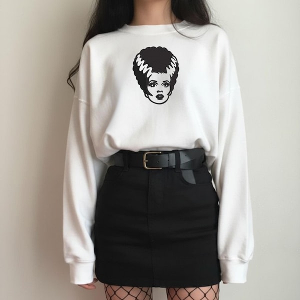 Könnte beinhalten: Ein weißes Langarm-Sweatshirt mit einem schwarzen Grafikdruck der Braut von Frankenstein auf der Brust, kombiniert mit einem schwarzen Minirock, einem schwarzen Gürtel mit silberner Schnalle und einer schwarzen Netzstrumpfhose unter dem Rock.