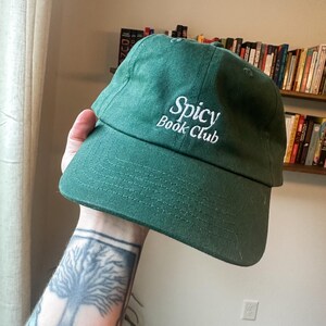 K&ouml;nnte beinhalten: Eine dunkelgr&uuml;ne Baseballkappe mit dem wei&szlig;en, gestickten Schriftzug "Spicy Book Club". Die Kappe hat einen gebogenen Schirm und ist aus weichem Material gefertigt. Ein l&auml;ssiger Hut.