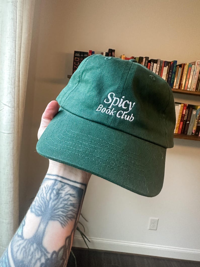 K&ouml;nnte beinhalten: Eine dunkelgr&uuml;ne Baseballkappe mit dem wei&szlig;en, gestickten Schriftzug "Spicy Book Club". Die Kappe hat einen gebogenen Schirm und ist aus weichem Material gefertigt. Ein l&auml;ssiger Hut.