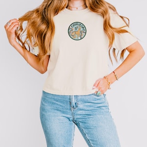 Op de afbeelding: Een crèmekleurig T-shirt met korte mouwen en een ronde afbeelding op de borst. De afbeelding toont een beige Steenbok-illustratie omringd door blauwe en grijze bloemelementen en sterren, met de tekst CAPRICORN in een boog aan de bovenkant.