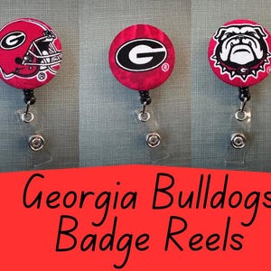 May include: Three red and white Georgia Bulldogs badge reels. One features a football helmet, one features the team logo, and one features a bulldog.