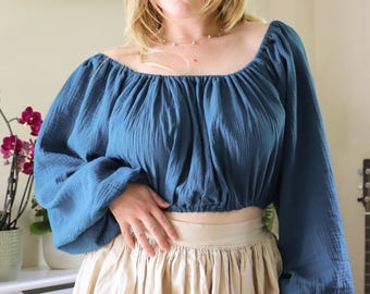 May include: A dark teal blue long-sleeved crop top featuring a wide, gathered square neckline and voluminous sleeves with elasticized cuffs. The garment is made of a textured, crinkled fabric and has an elasticized hemline that sits at the waist.