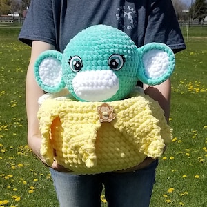 May include: A teal blue crocheted monkey plush toy with white eyes and a white crocheted face mask. The monkey is holding a yellow crocheted blanket with a small wooden monkey charm.