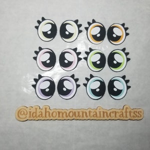 May include: Twelve colorful googly eyes with black pupils and white scleras. The eyes are arranged in three rows of four. Each eye has a different colored rim, including yellow, orange, pink, purple, green, and blue. The eyes are on a white background. @idaho mountaincrafts