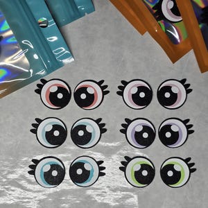 May include: A collection of cartoon eye stickers in various colors, including pink, blue, purple, and green, with black pupils and eyelashes. Some stickers are packaged in teal and orange holographic bags.