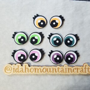 May include: Assortment of felt googly eyes with different colored pupils, including green, teal, purple, pink, and orange. The eyes are arranged in rows on a light brown wooden surface with the text "@idahomountaincraftss" printed on it.