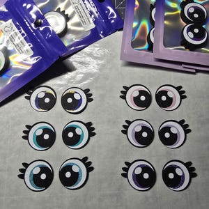 May include: A collection of felt eyes with lashes in various colors, including blue, pink, and purple, arranged on a light surface. Some eyes are packaged in clear plastic bags with text that reads "Idaho Mountain Crafts" and "25mm Favorits-Fancy Felt Eyes with Lashes."