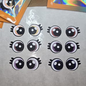May include: A collection of eye-shaped stickers with glitter accents in various colors, including red, gold, purple, and blue. Each sticker features a black pupil and a crescent moon design. The stickers are arranged on a white surface.