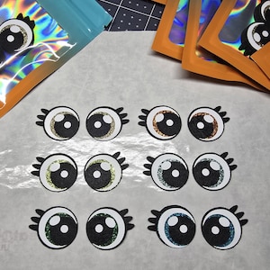 May include: A collection of decorative eye cutouts with glitter accents in various colors, including gold, brown, green, and blue. Each eye has a black pupil and white highlights, with black eyelashes. The cutouts are arranged on a light-colored surface.