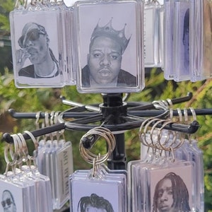 Könnte beinhalten: Eine Präsentation von durchsichtigen Schlüsselanhängern aus Kunststoff mit Schwarzweißbildern verschiedener Rapper. Die Schlüsselanhänger hängen an einem schwarzen Metallständer.