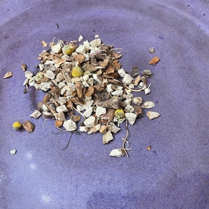 May include: A close-up of a pile of dried herbs and flowers on a purple surface. The herbs are a mix of light brown, tan, and white pieces, with some small yellow flower buds.