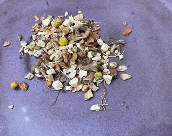 May include: A close-up of a pile of dried herbs and flowers on a purple surface. The herbs are a mix of light brown, tan, and white pieces, with some small yellow flower buds.
