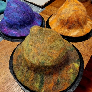 May include: Three felt hats in different colors and patterns. The hats are on a wooden surface. The hats are all pointed and have a wide brim.
