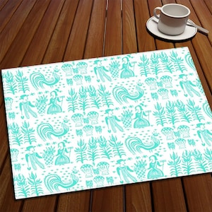 Pode incluir: Um jogo americano retangular branco com um padrão repetido de galos, figuras e elementos florais em azul-turquesa. O jogo americano está sobre uma mesa de madeira, com uma chávena de café e um pires no fundo.
