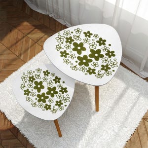 以下が含まれることがあります： オリーブグリーンの花柄が施された、白の腎臓型のサイドテーブル2つ。テーブルは明るい茶色の木製の脚が付いています。テーブルは白いふわふわのラグの上に置かれています。リビングルーム用の装飾的なテーブルです。