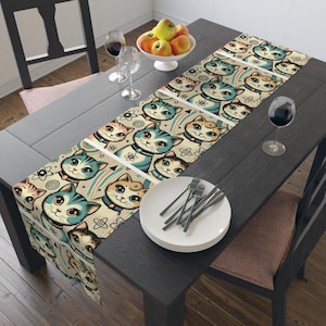 May include: A beige table runner with a repeating pattern of cartoon cats wearing space helmets and atomic symbols. The cats are in shades of blue, green, orange, and yellow. The runner is on a dark wood table with a white plate and silverware.