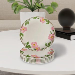 May include: A set of six white ceramic plates with a floral design. Each plate features pink roses and green leaves around the rim. The plates have a scalloped edge and a gold trim. A decorative and elegant tableware set.