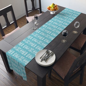 Peut inclure: Un chemin de table turquoise avec un motif géométrique blanc. Le chemin de table est posé sur une table en bois foncé avec deux chaises et un bol de fruits.