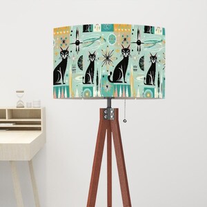 May include: A floor lamp with a tripod base and a drum shade featuring a retro-style pattern of black cats and geometric shapes on a teal background.