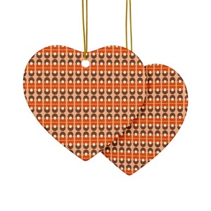 Op de afbeelding: Twee hartvormige ornamenten met een retro patroon van bruine, oranje en crèmekleurige vormen op een perzikachtergrond. De ornamenten hangen aan goudkleurige touwtjes, perfect voor vakantiedecoratie.