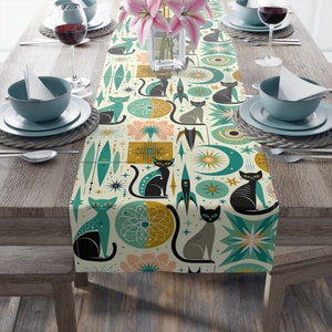 May include: A cream-colored table runner with a mid-century modern design featuring black cat silhouettes, rockets, and starbursts. The runner is decorated with teal, gold, and pink geometric shapes. The runner is placed on a wooden table set for a meal.