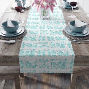 Peut inclure: Un chemin de table turquoise et blanc avec un motif répété de figures stylisées, de fleurs et d'oiseaux. Le chemin de table est posé sur une table en bois avec des couverts et un vase de fleurs roses.