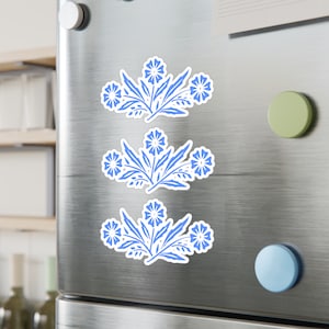 May include: Three blue floral designs on a silver refrigerator. Each design features a cluster of stylised flowers with leaves.
