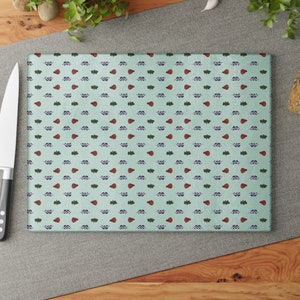 May include: A light blue cutting board with a repeating pattern of small red, green, and purple leaves. The board is made of glass and has a smooth surface.