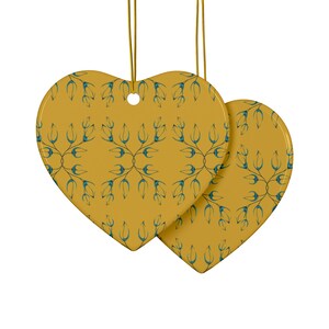Op de afbeelding: Twee gele hartvormige keramische ornamenten met een blauw bloemenpatroon. De ornamenten hangen aan gouden touwtjes.