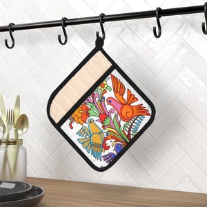 May include: A white oven mitt with a colourful floral and bird design. The design features orange, pink, yellow, blue, and green colours. The mitt is hanging on a black metal rack.