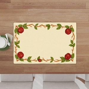 May include: Rectangular beige placemat with a border of red apples, green leaves, and brown branches. The placemat is on a wooden table, with a white plate and spoon visible in the corner. A decorative item for the dining table.