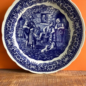 May include: A blue and white ceramic plate with a decorative border and a scene of people in a village setting. The scene includes a man playing a violin, a woman looking out a window, and a group of people gathered around a table.