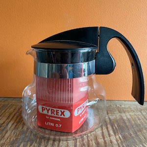 May include: A clear glass Pyrex teapot with a black handle and lid. The teapot has a silver band and a red box inside with the Pyrex logo and "Litri 0.7" printed on it. The teapot is on a wooden surface.
