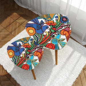 May include: Two white nesting tables with a colourful floral and bird design. The tables have a unique, leaf-shaped top and wooden legs.