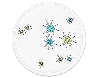 May include: A white, round coaster with a stitched edge. The coaster features a mid-century modern design of black starbursts with teal and yellow-green accents. The coaster is made of a textured material.